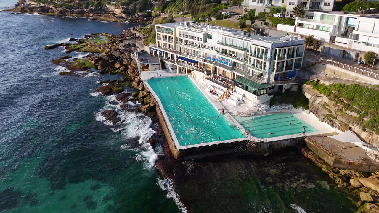Rotating aerial shot of an ocean pool at Bondi Beach, Australia, with waves crashing dramatically against the rock walls. Cinematic drone footage.