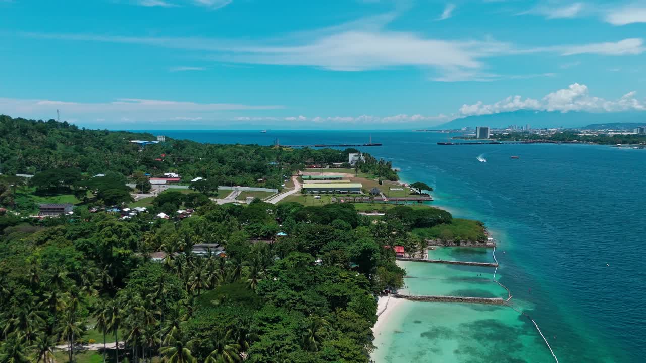 Stunning aerial drone shot of a tropical island coastline with turquoise waters, lush greenery, and distant city skyline. Perfect for travel, real estate, and lifestyle projects