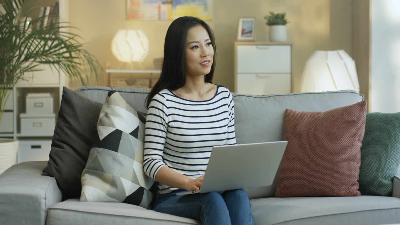 줄무늬 셔츠를 입은 젊은 여성은 랩탑에서 노트북 작업을 하고 거실에 있는 소파에 앉아 생각하고 있습니다 1