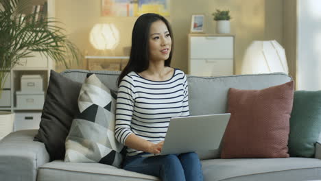 young woman in striped shirt working with laptop on laps and thinking sitting on the sofa in the living room 1