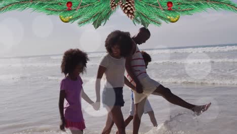 animation of christmas decorations over happy biracial parents and children walking on sunny beach