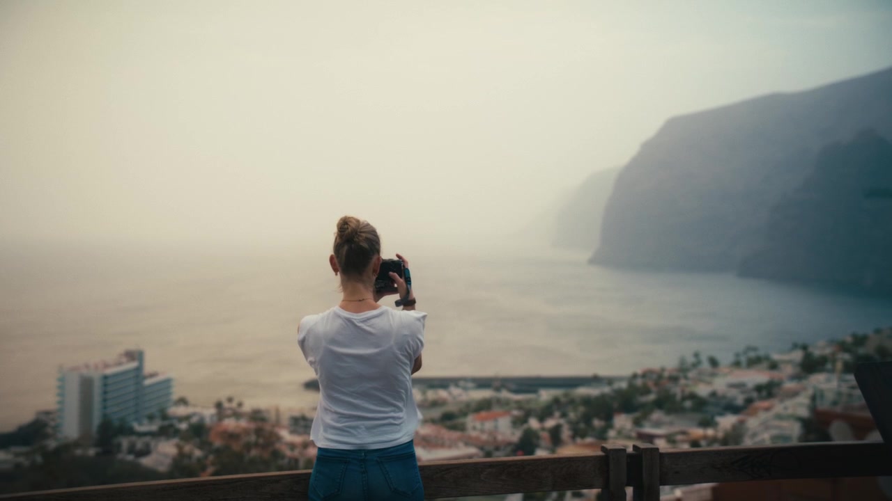 Woman photographing the coastal town and hazy sea cliffs of Los Gigantes, Tenerife, from an elevated viewpoint under soft, calima-diffused light.
