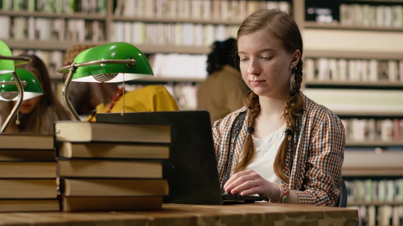 Ung student som jobber på bærbar PC, tar notater ved et biblioteksbord med en åpen bok