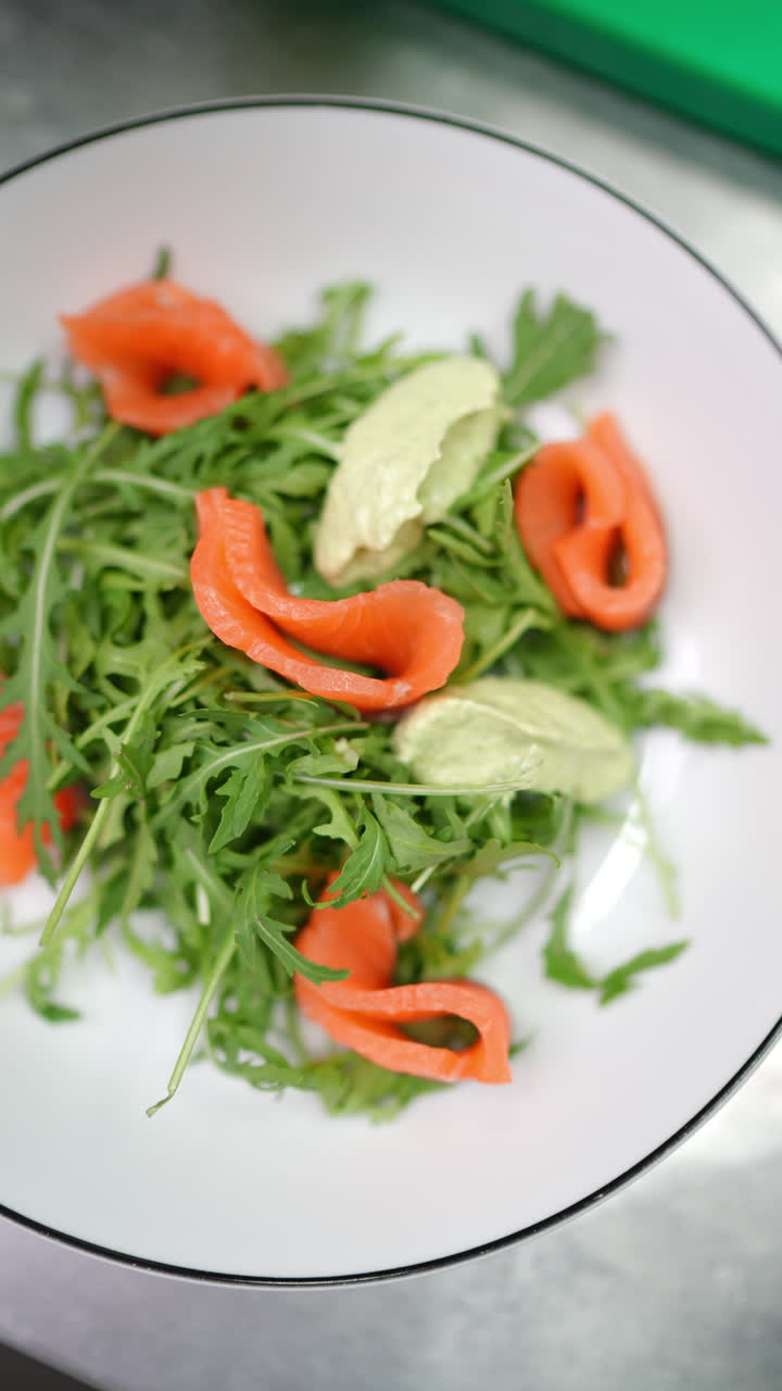Vertical footage Gourmet salmon and arugula salad receiving delicate wasabi paste garnish by professional chef inside pristine restaurant kitchen environment