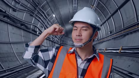close up of asian male engineer with safety helmet flexing his bicep and smiling to camera while standing in underground subway tunnel