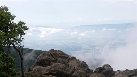 Una-Toma-Estática-Desde-La-Cima-De-Una-Montaña-Rocosa-Captura-Una-Vista-Impresionante-De-Una-Tierra-Brumosa-Debajo.