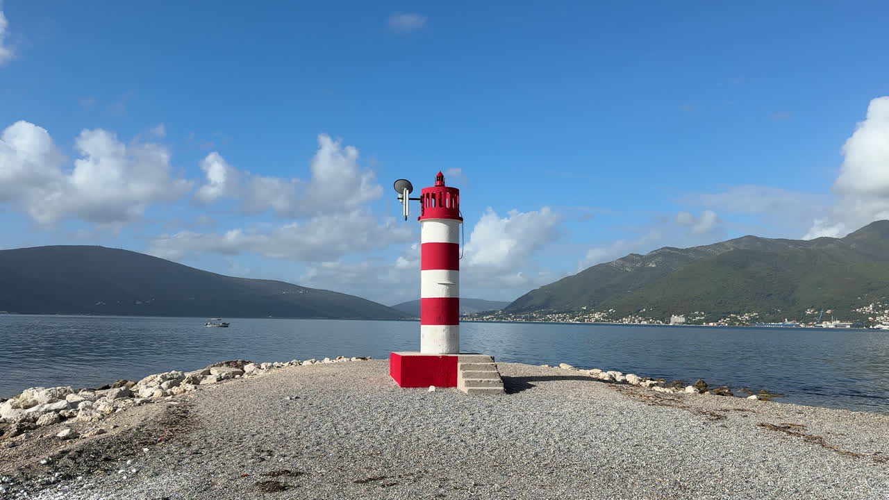 A red-and-white coastal lighthouse stands on a rocky shore under a clear blue sky, surrounded by calm sea and distant mountains. Perfect for travel, maritime, navigation and scenic landscape themes