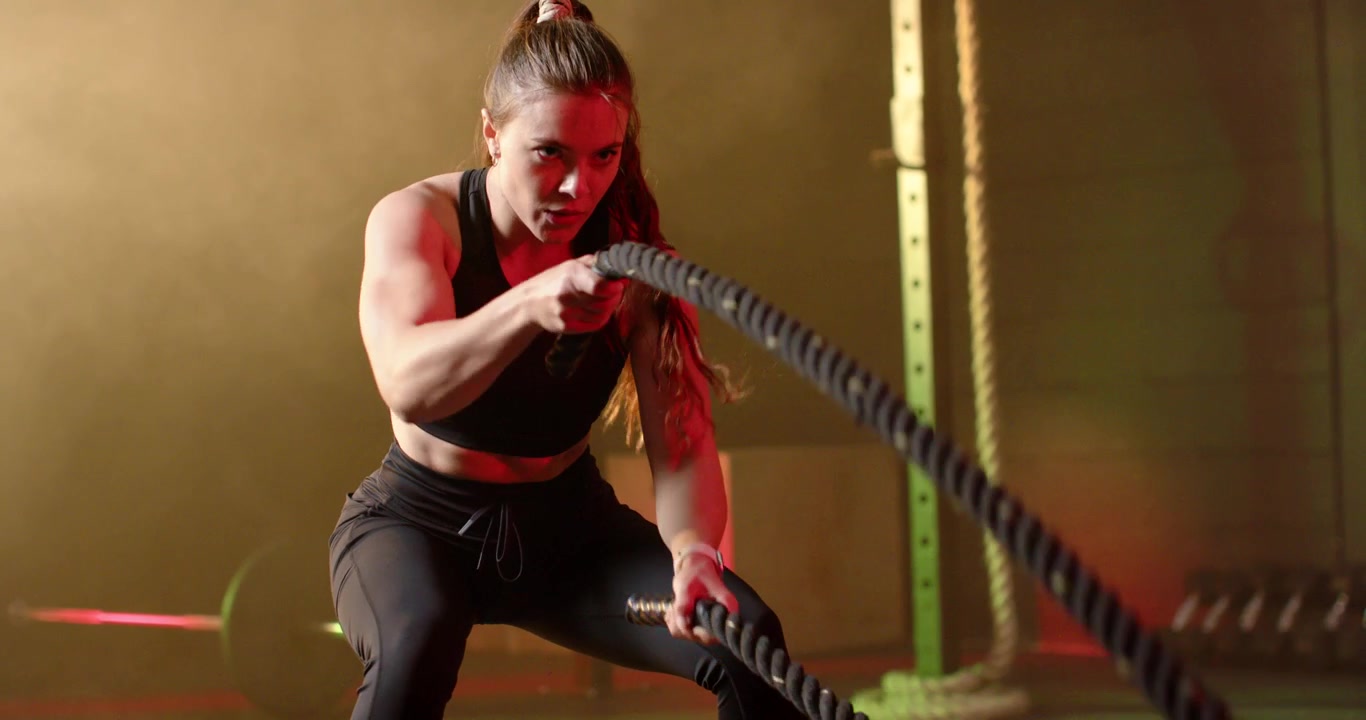Mujer con equipo negro haciendo cuerdas de batalla en el gimnasio, iniciando tirones para fuerza, sosteniéndolas y respirando