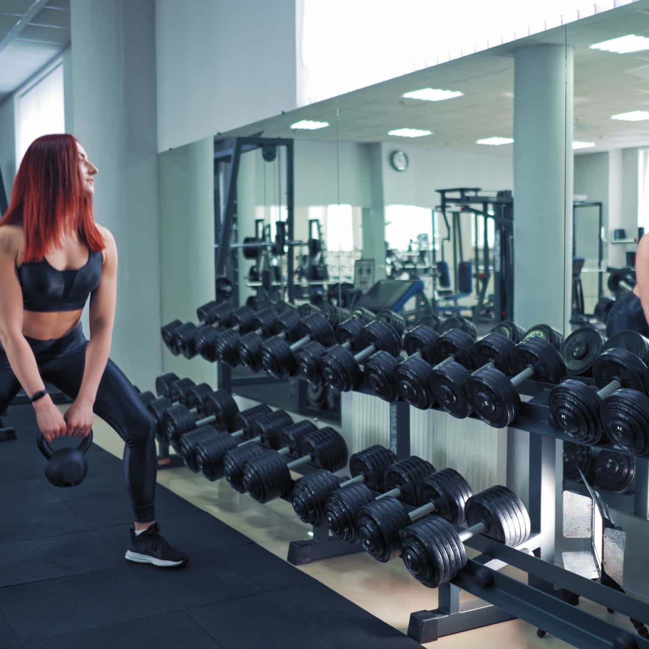 Attractive woman with kettle bell in the gym. Side view of slender female in sporty top and leggings doing workout. Strong red-haired girl lifting kettlebell while looking in the mirror.