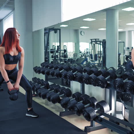 attractive woman with kettle bell in the gym. side view of slender female in sporty top and leggings doing workout. strong red-haired girl lifting kettlebell while looking in the mirror.