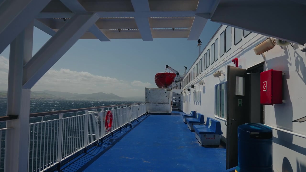Description:
Empty ferry deck with blue floor lifeboat and railing overlooking sea and mountains