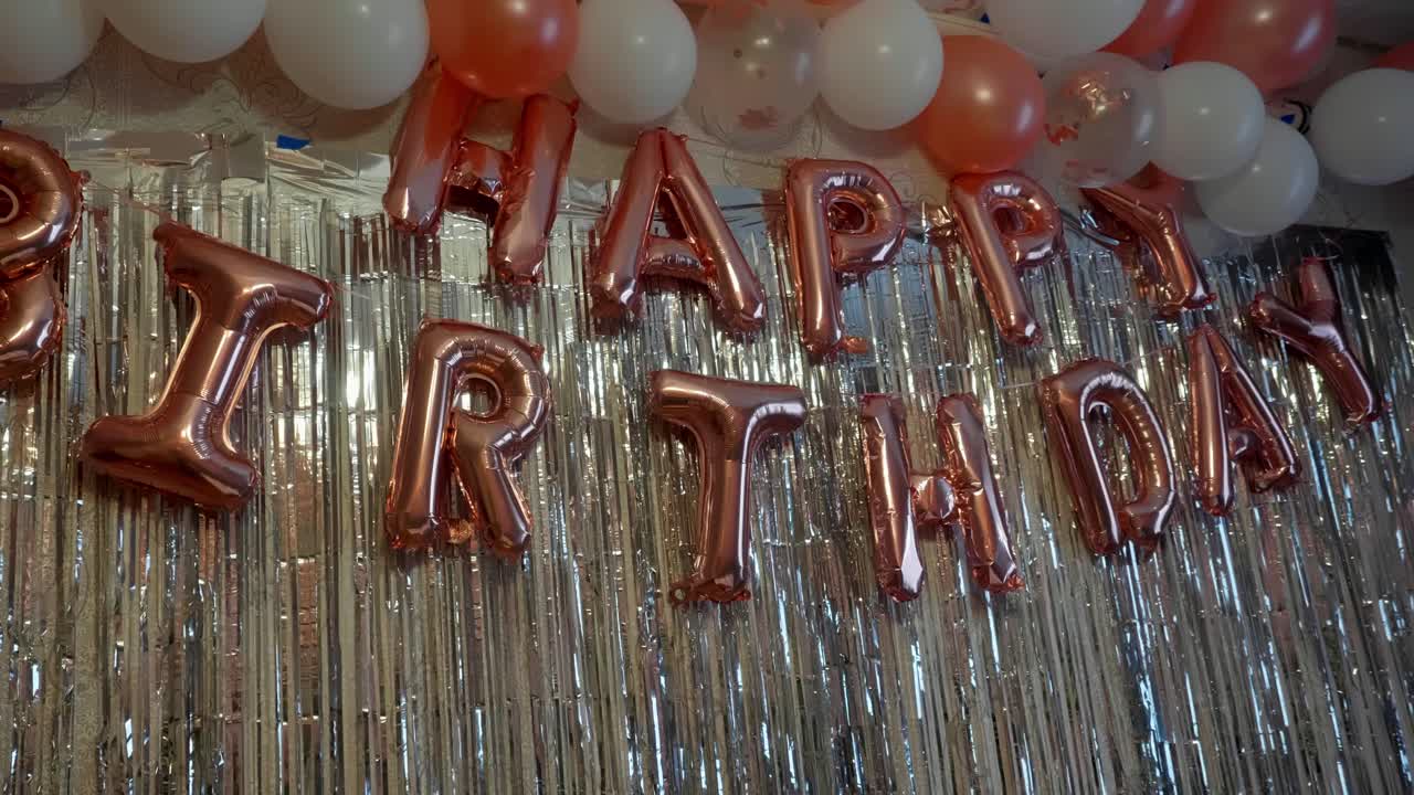 Indoor birthday party setup with metallic streamers, rose gold balloons spelling "Happy Birthday," and white and rose gold balloons above