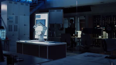 in industrial robot desing research laboratory: robotic arm prototype standing illuminated on the desk. in the dark background various high tech devices, computers, digital whiteboard and blueprints