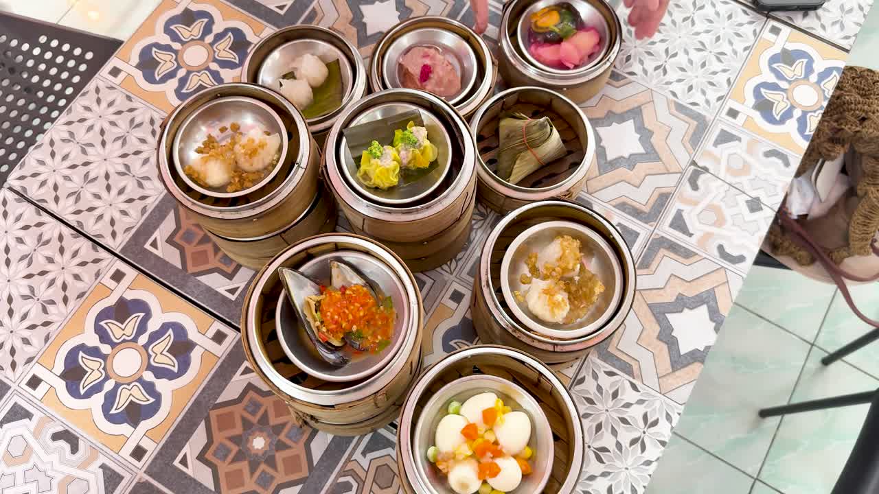 Hands arrange assorted dim sum baskets on patterned table, overhead view, bright natural lighting, Phuket