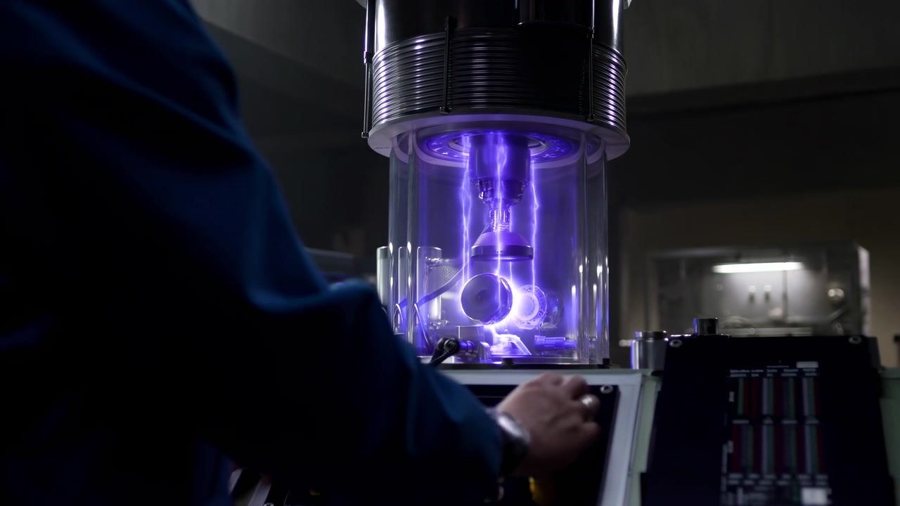 Hands operate control console beside clear cylindrical chamber where violet plasma arcs glow around metal fixtures in dim research laboratory, creating precise industrial light and technical