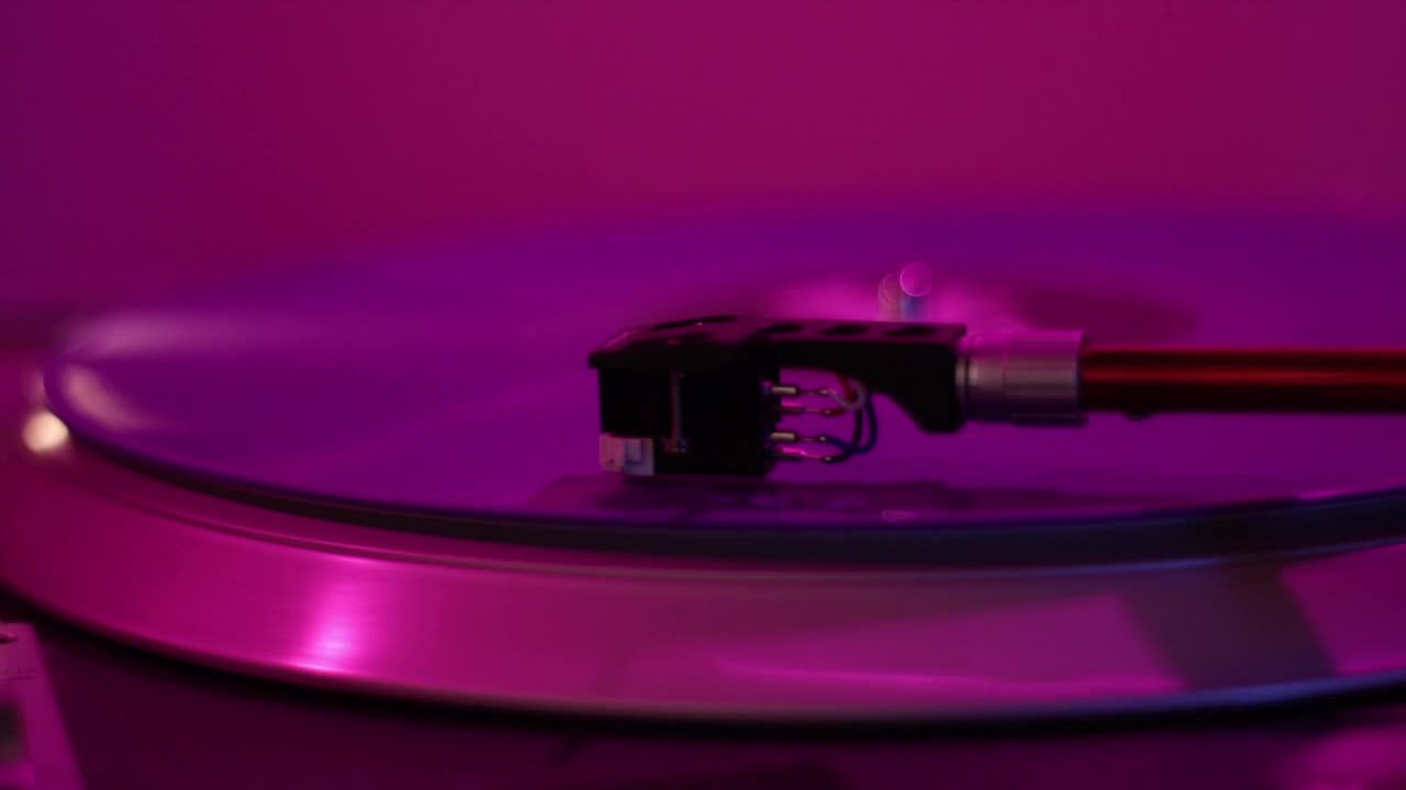 En pladespillernål snurrer under de lilla og lyserøde lys fra en DJ-opsætning med lys, lyd og røg. En DJ spinner den skæve plade. Starter og stopper pladespilleren