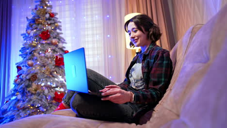 woman on laptop at holidays. a woman sits on a couch focused on her laptop, surrounded by festive decorations and a christmas tree in the evening
