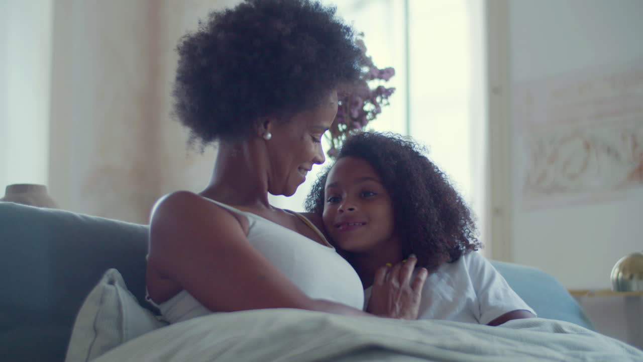 plano medio de la feliz madre afroamericana y su hija acostadas en la cama y hablando juntas