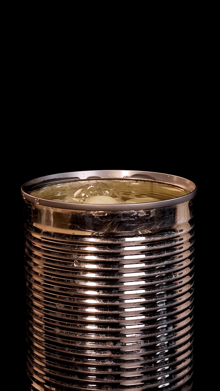 Una fruta de longan pelada se deja caer en una lata de metal abierta llena de almíbar, creando un salpicadura. Iluminación de estudio, fondo negro, cámara estática.