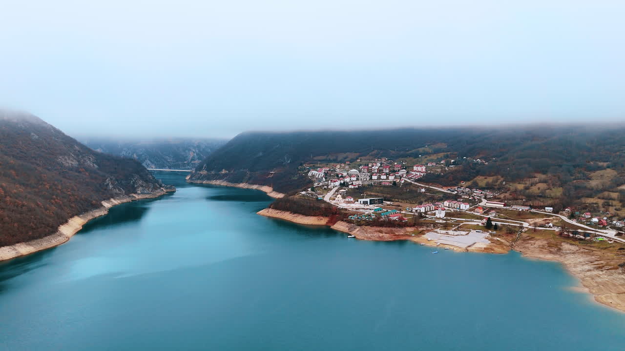Аэропанорама поселка Плужине и водохранилища Пивского озера. Широкая перспектива города на склоне холма, откуда открывается вид на бирюзовые воды Пивского озера под туманным небом.