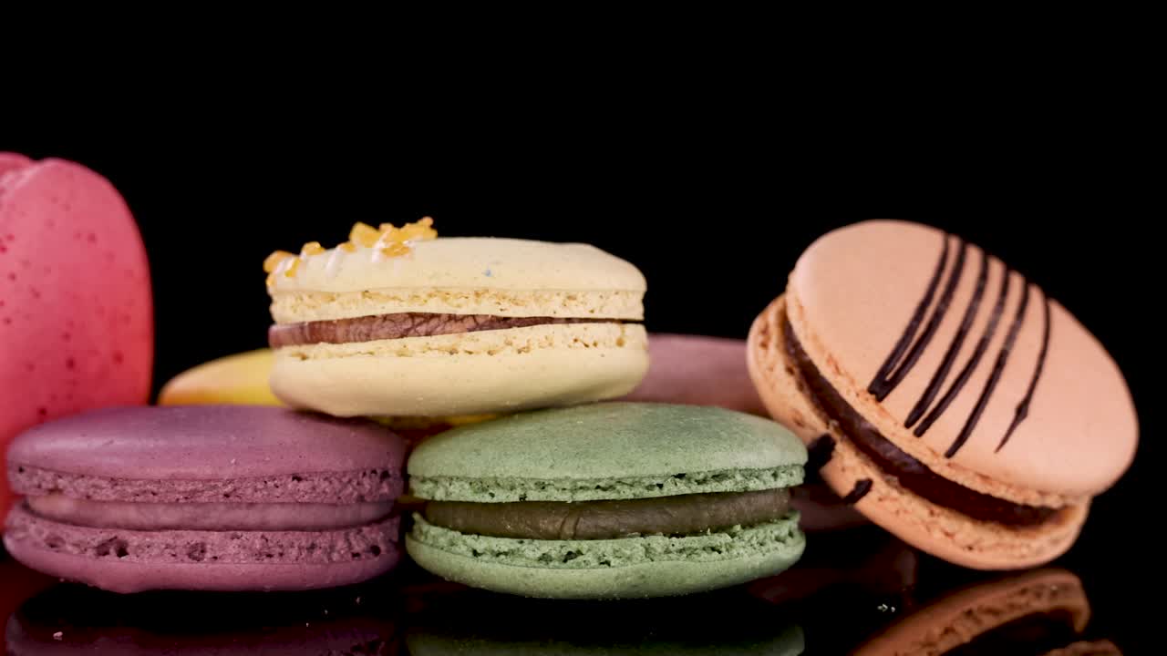 Colorful assorted macarons rotate on glossy black surface under dramatic studio lighting, close-up view