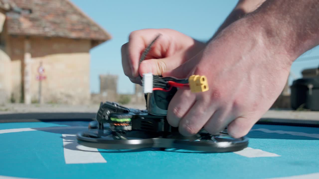 Detailed view of a pilot plugging the yellow power connector into an FPV drone battery with a blurred building in the background