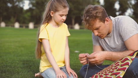 father fixing kite sitting on meadow with his little daughter