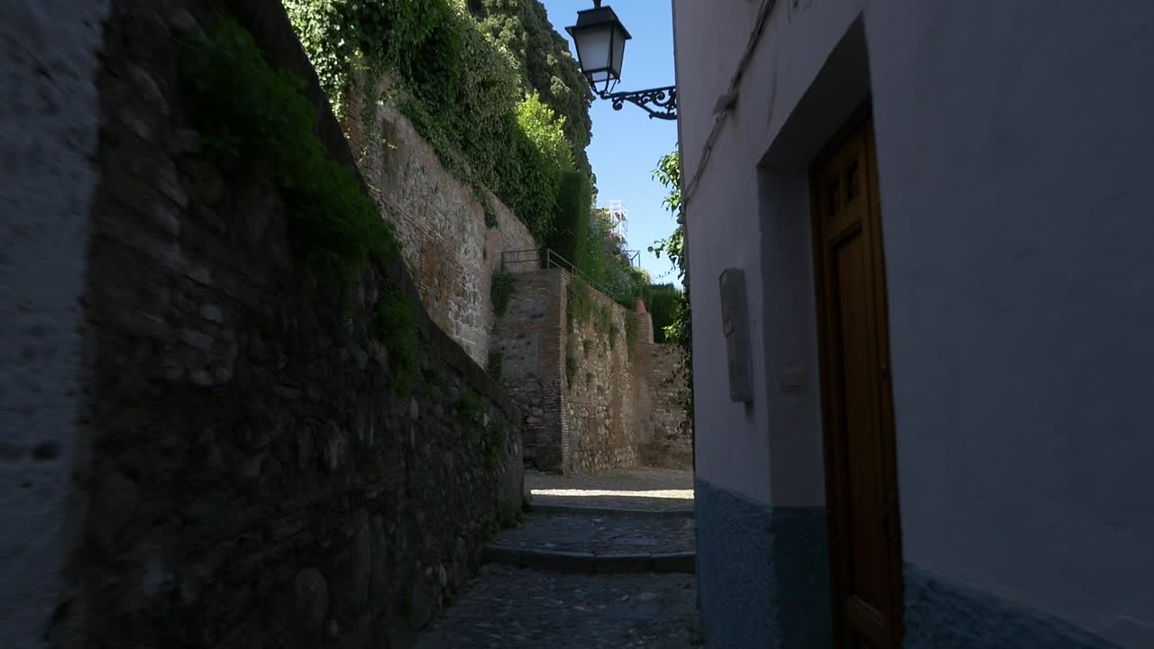 Un'inquadratura dal basso di una passeggiata in uno stretto vicolo in salita con mura in pietra e un tradizionale lampione in ferro battuto. Autentica atmosfera mediterranea nello storico quartiere dell'Albaicín.