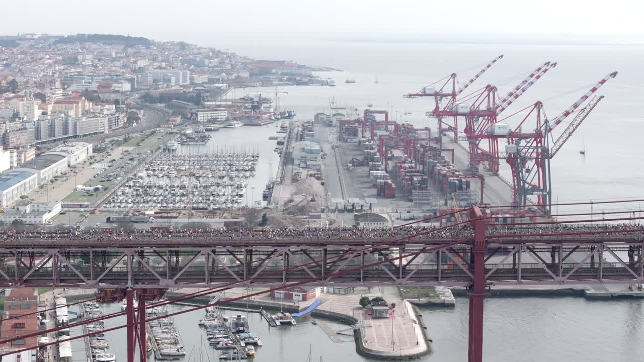 Side-on aerial drone shot of half marathon and 10k runners crossing the iconic red 25th April suspension bridge above the Lisbon waterfront, marina, port docks, and industrial cranes in Portugal