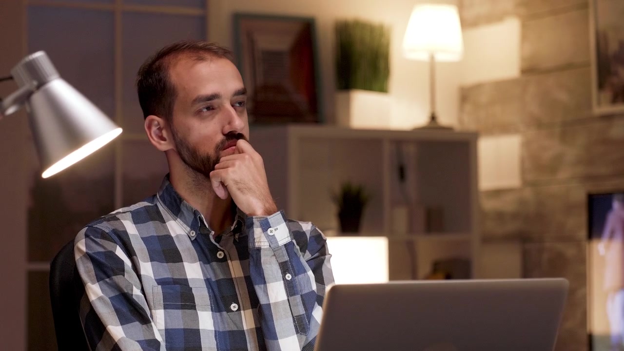 Thoughtful Man Working on Laptop in Home Office at Night