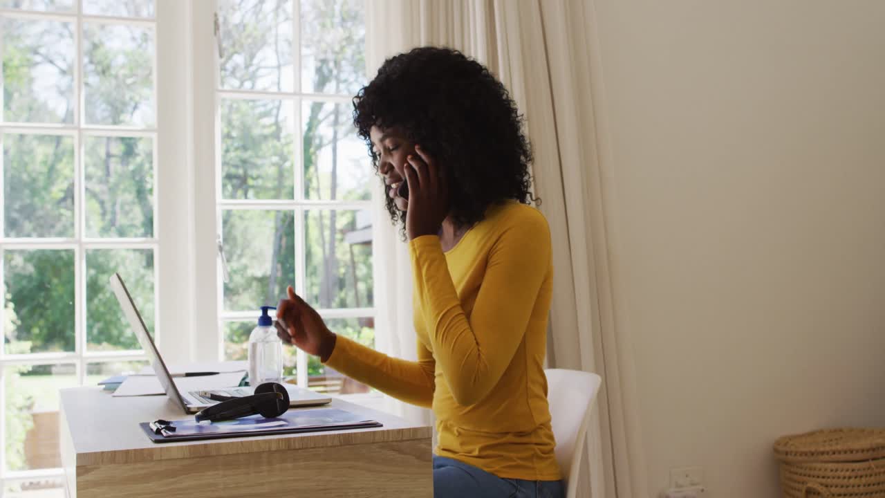 mujer afroamericana usando una computadora portátil y hablando en un teléfono inteligente mientras está sentada en su escritorio en casa