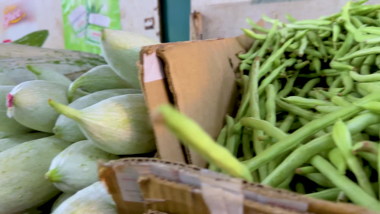 Close-up tracking shot of bitter melon, cucumbers, and beans in bright indoor market lighting