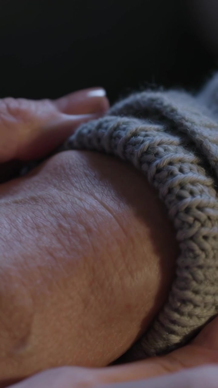 Video verticale: Mani che afferrano il bordo del polsino, arrotolando un polsino di maglia grigio in una stanza in penombra, rivelando il polso.