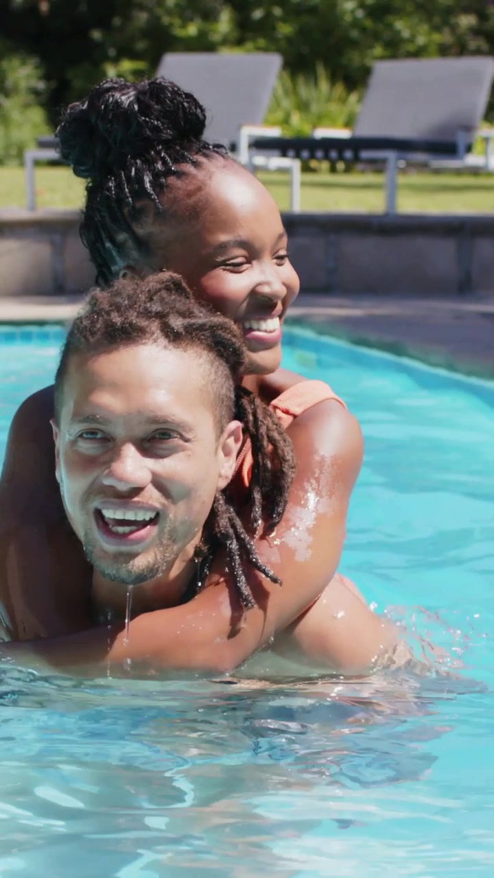 Video verticale: Uomo che porta in braccio una donna in bikini color corallo che gli sale sulla schiena in piscina nel cortile, giocosamente.