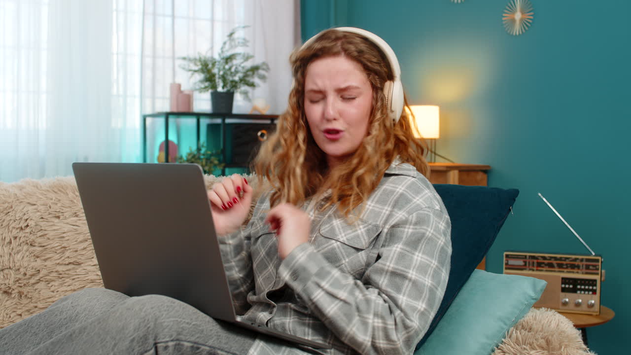 Woman sits on sofa wearing headphones focused looking at laptop during video call or online course