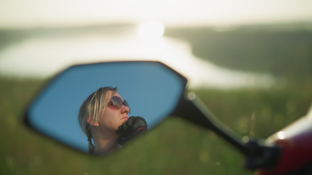 A close-up mirror reflection shows a focused woman wearing sunglasses, gently touching her face while gazing into the distance, the background reveals a blurred field and water body