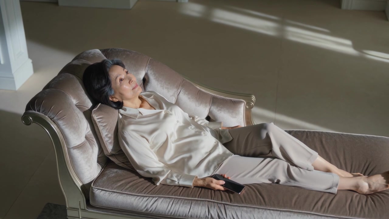 Anziana donna asiatica a riposo, che si muove leggermente su una chaise longue a casa, accarezzata dalla luce del sole, mentre tiene un telecomando e indossa seta.