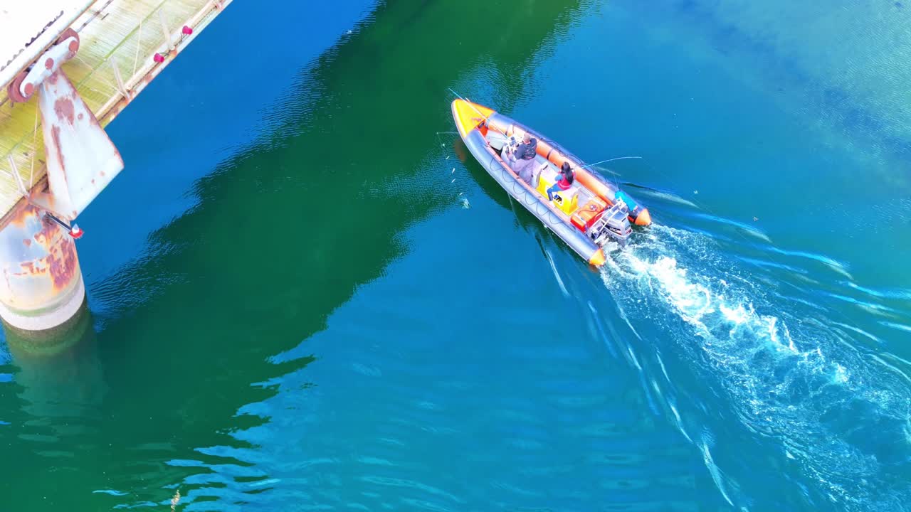 Top-down aerial view of a small inflatable boat navigating under a broken bridge. Calm water reflections and serene motion in a harbour setting