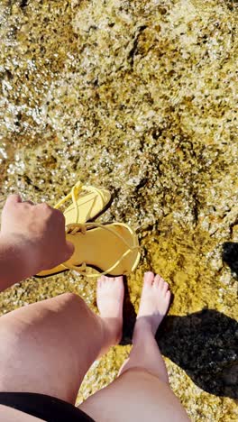 stepping barefoot into clear shallow water with sandals in hand