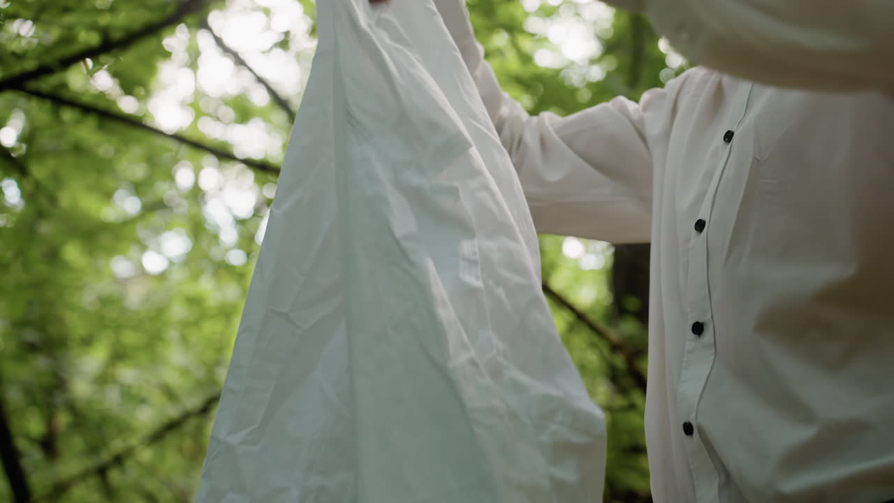 Close view of man in white removing coat in forest surrounded by blurred green leaves and soft natural light, highlighting outdoor moment, peaceful woodland setting, and lifestyle detail