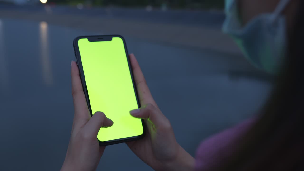 tiro de mano: mujer turista sosteniendo un teléfono de pantalla verde por la noche y una mujer usando una máscara para prevenir enfermedades.