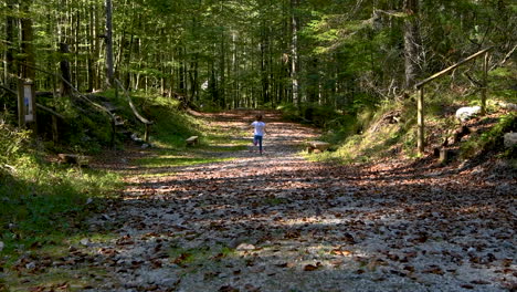 Girl-running-in-forest-behind-mother,-playing-in-woods,-foothpath-with-autumn-foliage,-follow-zoom,-shot-from-behind,-facing-away