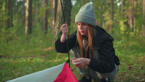 woman squats outdoors carefully preparing tent for camping as she holds flexible pole and attaches fabric, sunlight glows warmly over her focused face while she works in lush forest surroundings