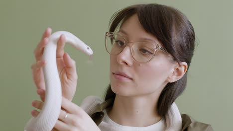 portrait of a young woman with eyeglasses holding a white pet snake around her neck and hands on a green background 2