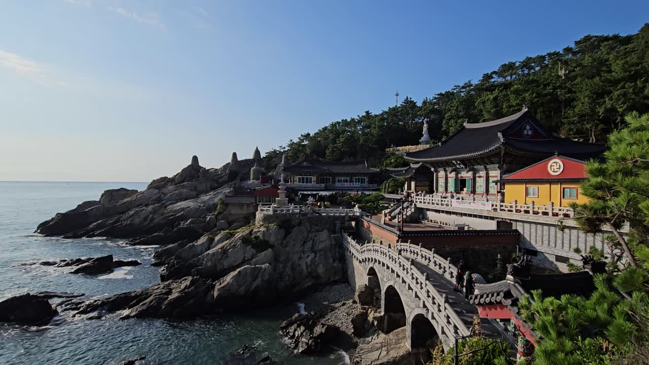 Haedong Yonggungsa Temple with bridge crossing over to grounds in the morning as ocean waves crash on cliffs below, Tourists throw coins for luck -Busan, South Korea - establishing shot