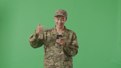 asian man soldier smiling and pointing to smartphone in his hand while standing in the green screen background studio