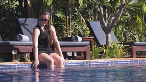 long shot of a young woman sat at the side of a tropical hotel pool with a refreshing cocktail