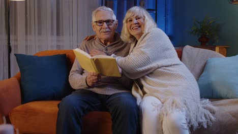 senior grandparents couple relaxing reading book talking enjoying leisure hobbies at night home