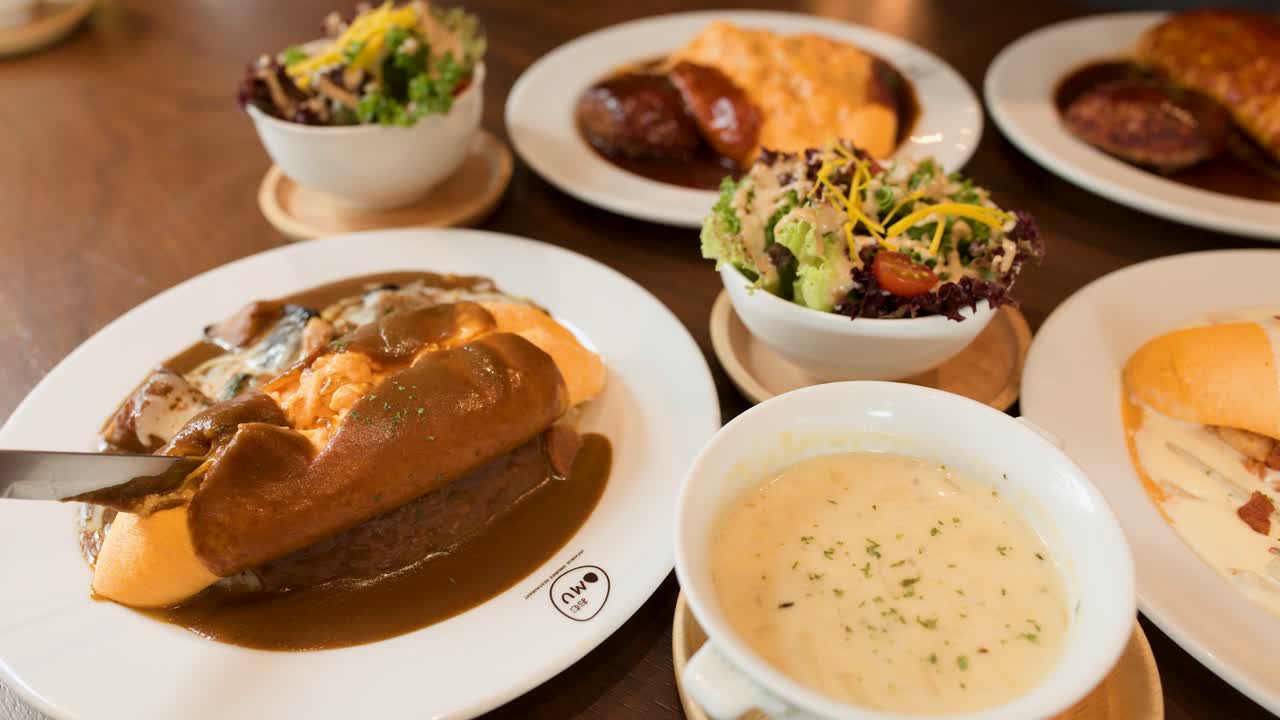 Hand slices omelette rice with brown sauce, surrounded by soup, salad, bread, and warm lighting