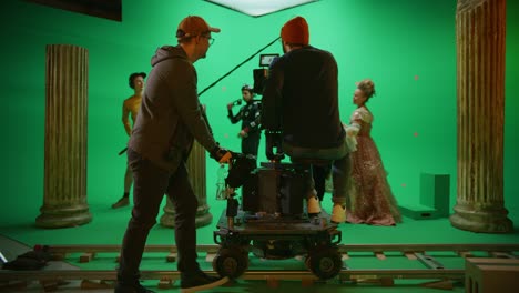 on film studio set shooting history movie green screen scene. moving cameraman on railway trolley shooting two costumed actors while director controls process. crew on big budget filmmaking